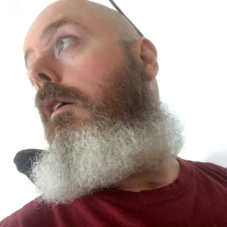 A selfie, taken from a low angle and straight on of me looking over my right shoulder, wearing a red t-shirt and a mid-length, largely greying beard.